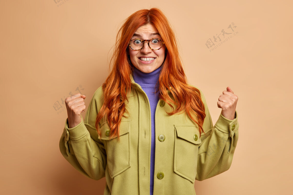 人兴奋快乐的红发女人握紧拳头 微笑着 期待着听到一些可怕的穿着绿色夹克的声音站立佩戴快乐