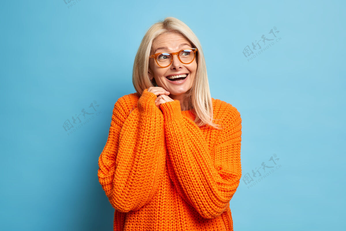 乐观快乐的金发高级欧洲女人回忆一些愉快的事情快乐地笑着 双手紧握着戴着眼镜宽松的针织橙色套头衫编织女性老年人