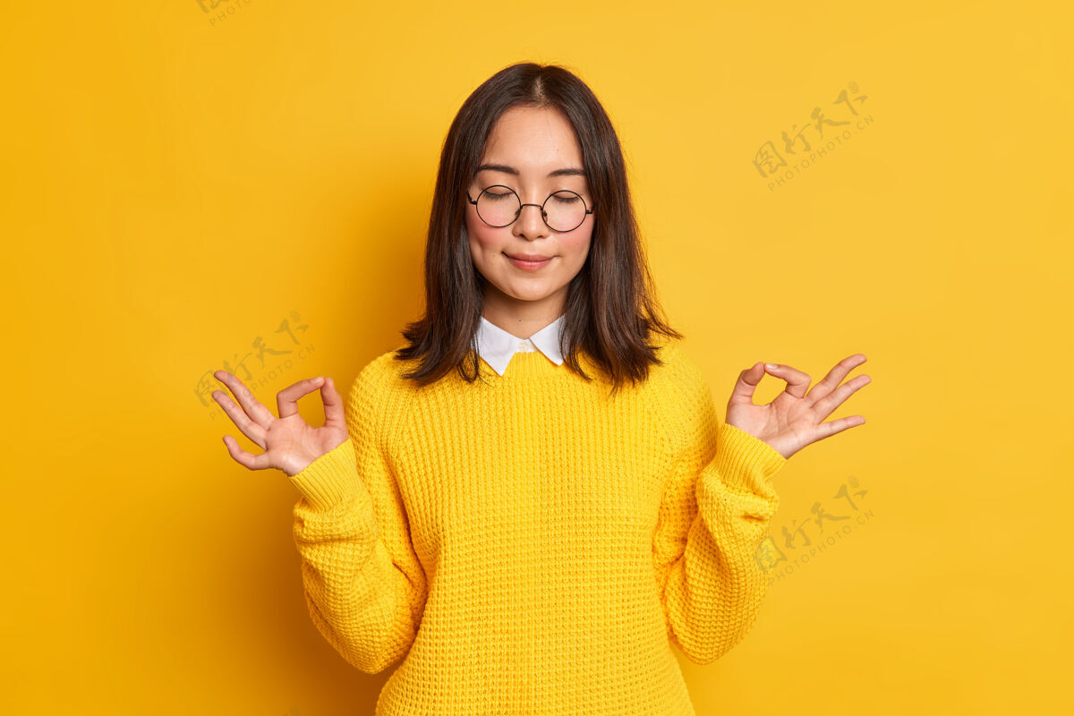 可爱冷静的亚洲年轻女子展示禅宗或OK标志冥想心情好站着闭着眼睛穿着休闲毛衣和眼镜姿势黄色眼镜
