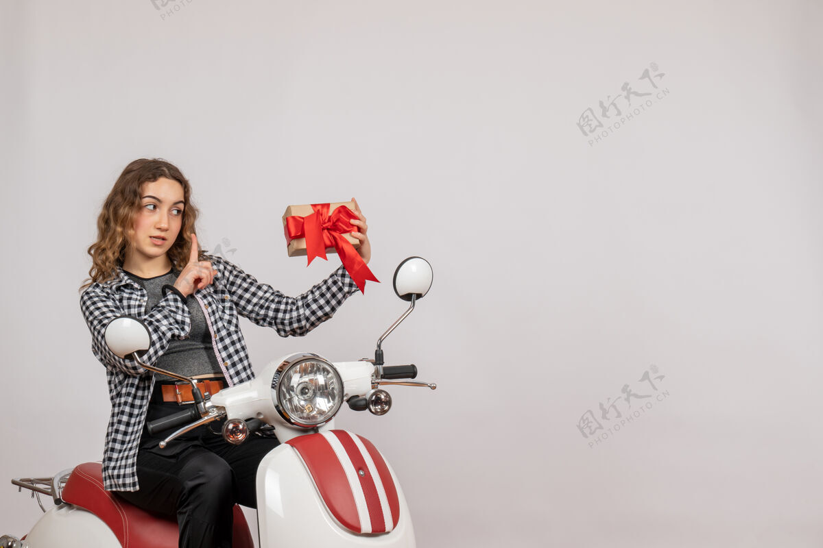 车辆美丽的年轻女子骑着轻便摩托车 拿着礼物站在灰色的墙上美丽的年轻女子摩托车摇滚