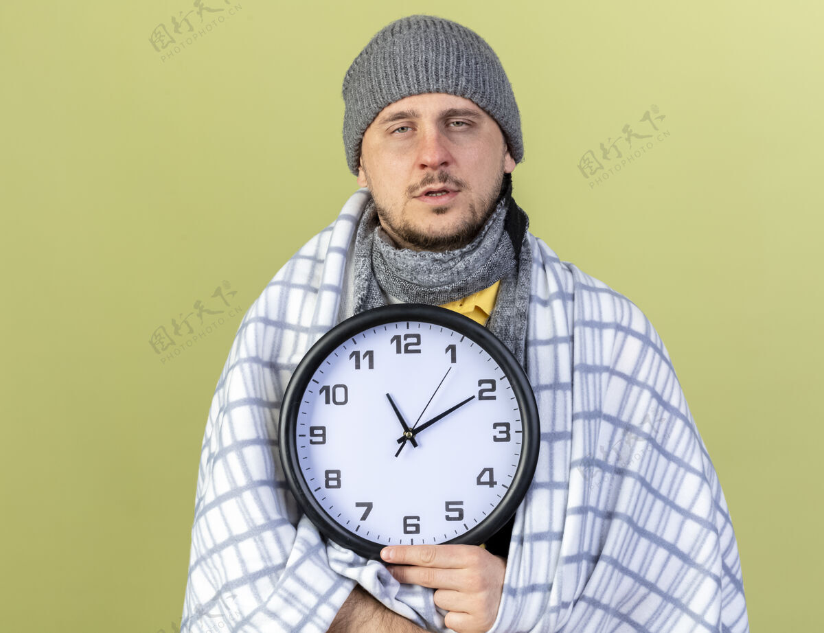 苍白苍白的年轻金发病斯拉夫男子戴冬季帽子和围巾包裹在格子布举行的橄榄绿墙上的复制空间隔离时钟格子围巾裹