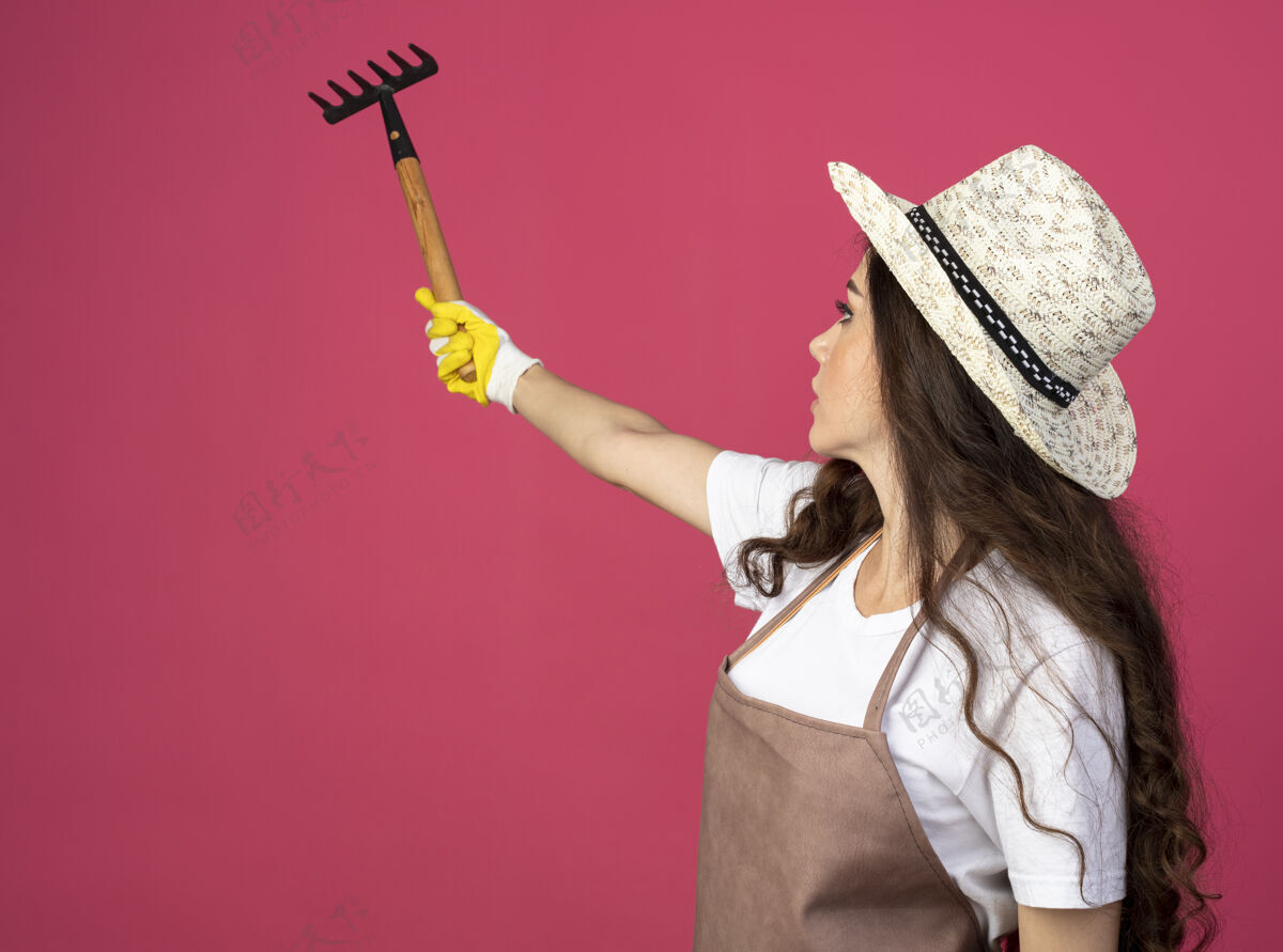 穿自信的年轻女园丁穿着制服 戴着园艺帽和手套 拿着并看着隔离在粉色墙壁上的耙子 还有复制空间持有花园年轻