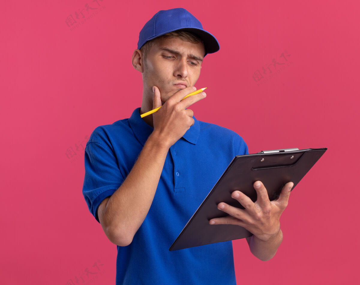 铅笔困惑的年轻金发送货男孩把手放在下巴上拿着铅笔 看着粉红色的剪贴板年轻交货粉红色