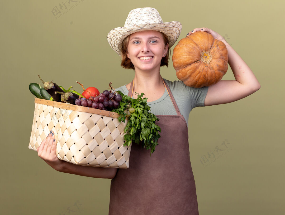 南瓜微笑着的年轻的斯拉夫女园丁戴着园艺帽 肩上扛着蔬菜篮子和南瓜 放在橄榄绿上肩膀蔬菜斯拉夫