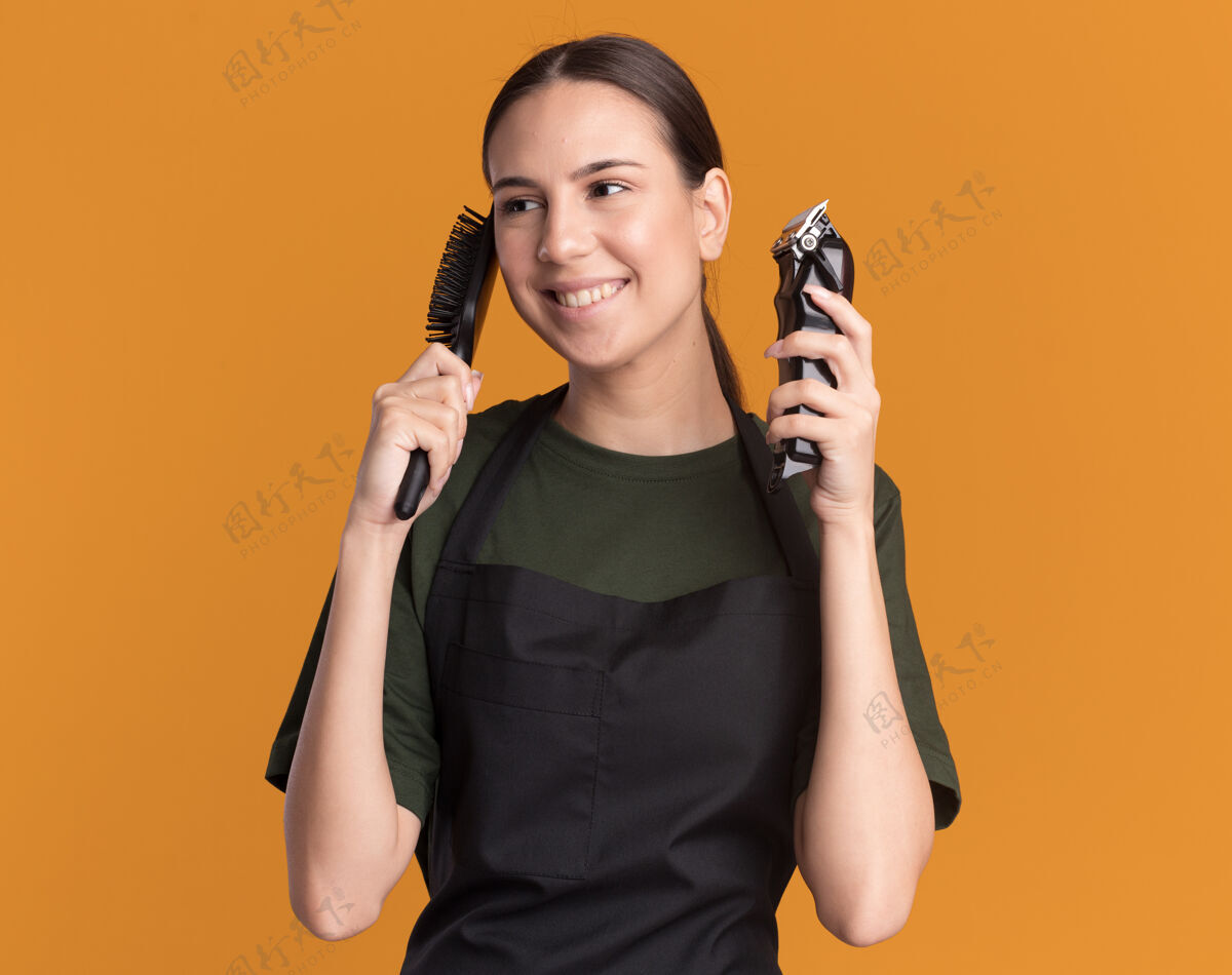 年轻人身着制服的年轻黑发理发女拿着理发器和梳子看着橙色的一边剪子梳子请拿着