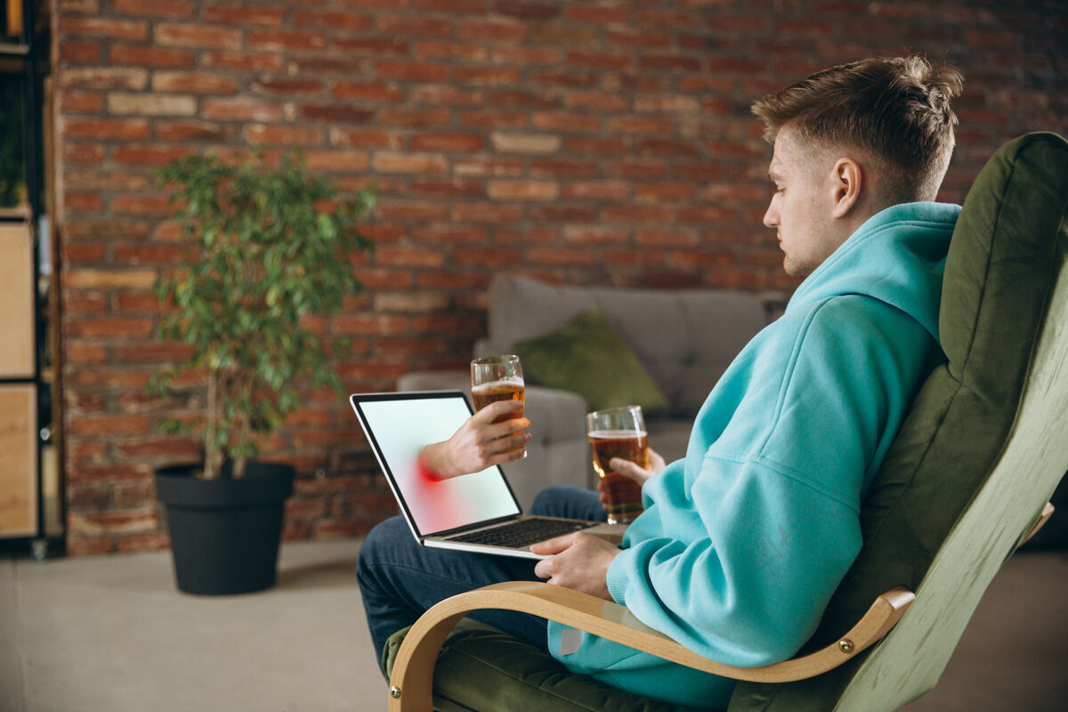 病毒年轻人在虚拟视频通话中与朋友见面时喝啤酒远程在线会议 在家里用笔记本电脑聊天远程安全会议和娱乐的概念周末室内玻璃