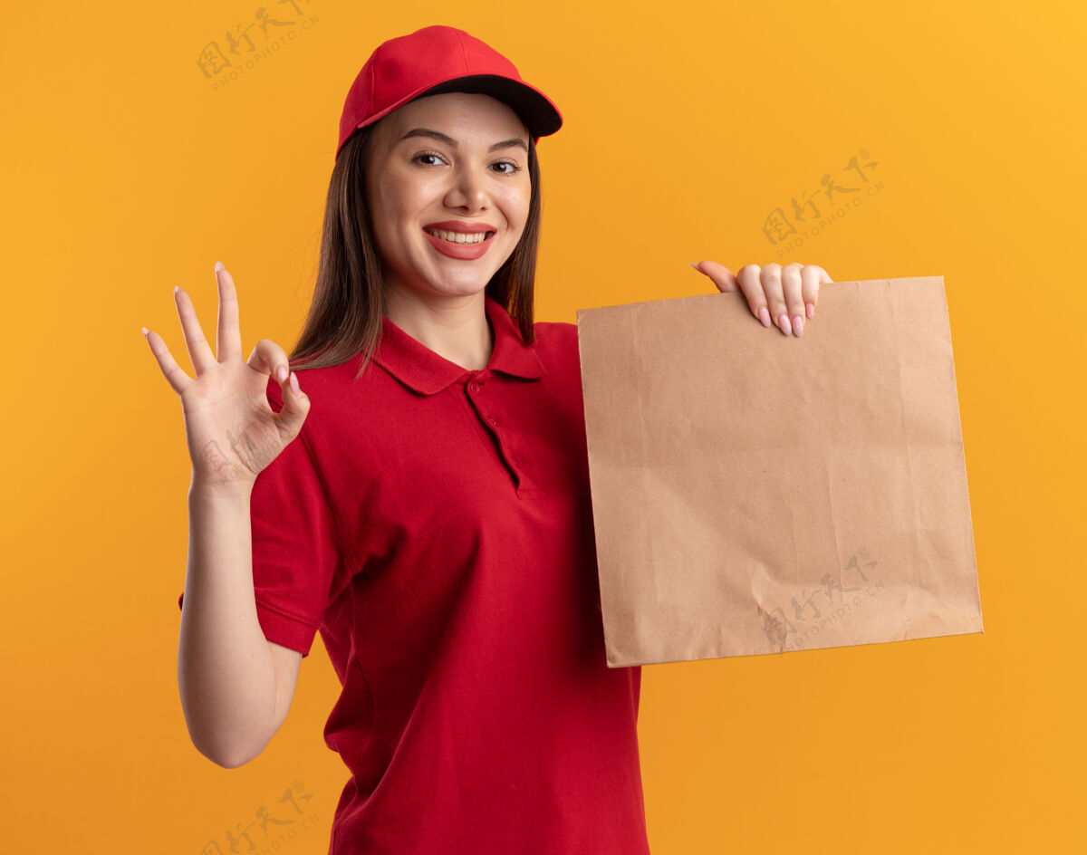 包装穿着制服的微笑漂亮的女送货员拿着纸包裹 在橙色上做着手势“ok”手势橙色手势标志