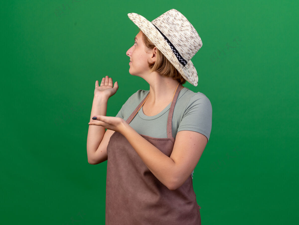 园艺高兴的年轻斯拉夫女园丁戴着园艺帽 看着并指着绿色的背面背部花园指点