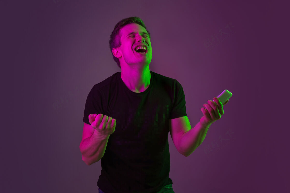 情感使用智能手机 下注 赢球霓虹灯下紫色工作室背景上的白种人肖像穿着黑色衬衫的漂亮男模人类情感的概念 面部表情 销售 广告男性成功工作