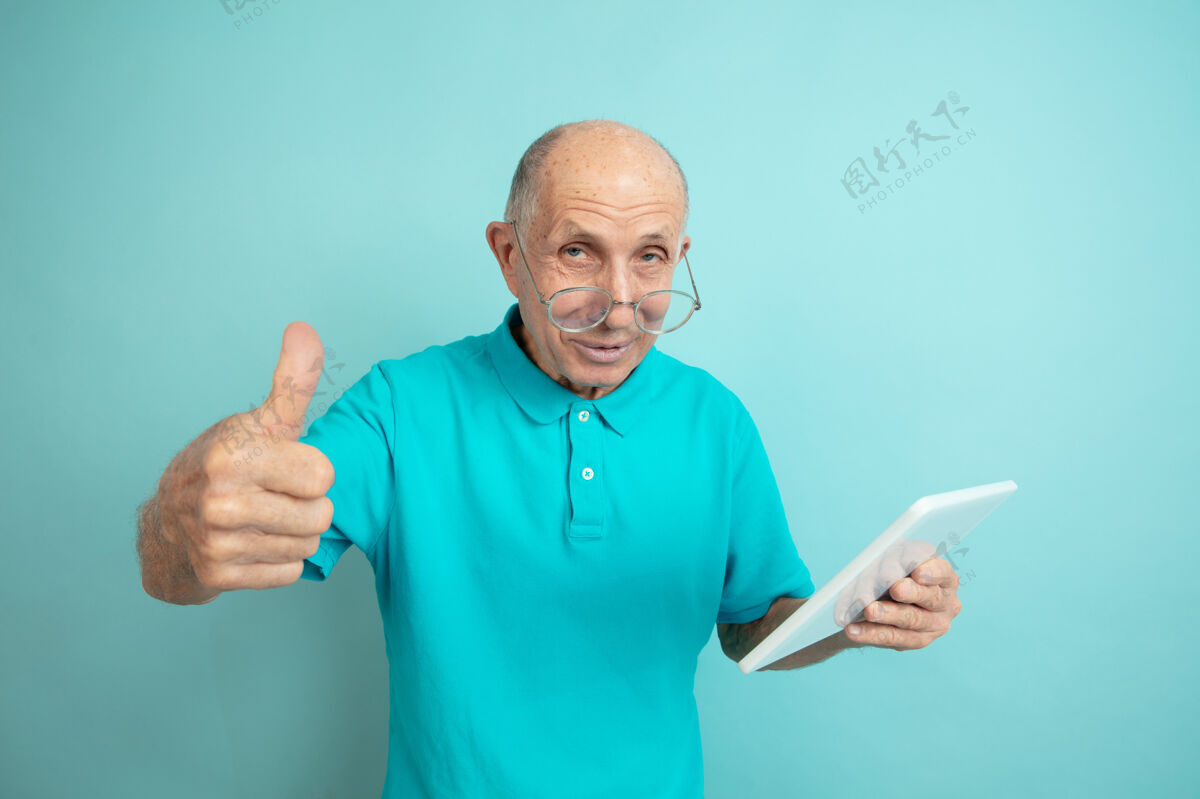 面部使用平板电脑 竖起大拇指蓝色工作室背景上的高加索老人肖像美丽的男性情感模型人类情感的概念 面部表情 销售 福利 广告空间长度英俊工作