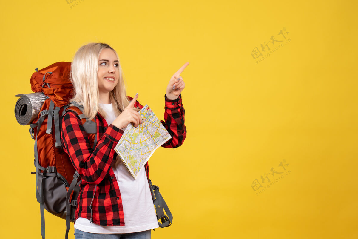 脸正面图快乐的金发女人背着她的背包拿着地图站在黄色的墙上手持站立前面