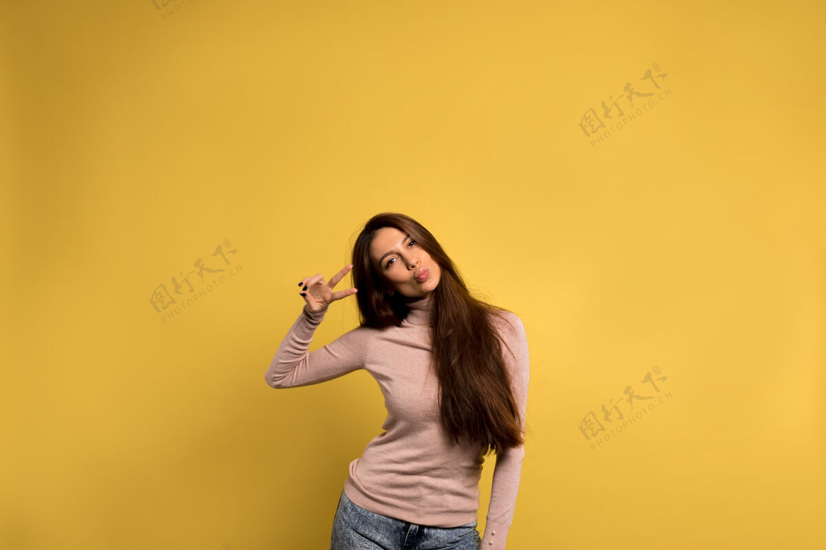 快乐有趣友好的女孩 棕色长发 穿着牛仔裤和粉色衬衫 在黄色的墙上送上一个吻和和平的标志姿态自我积极