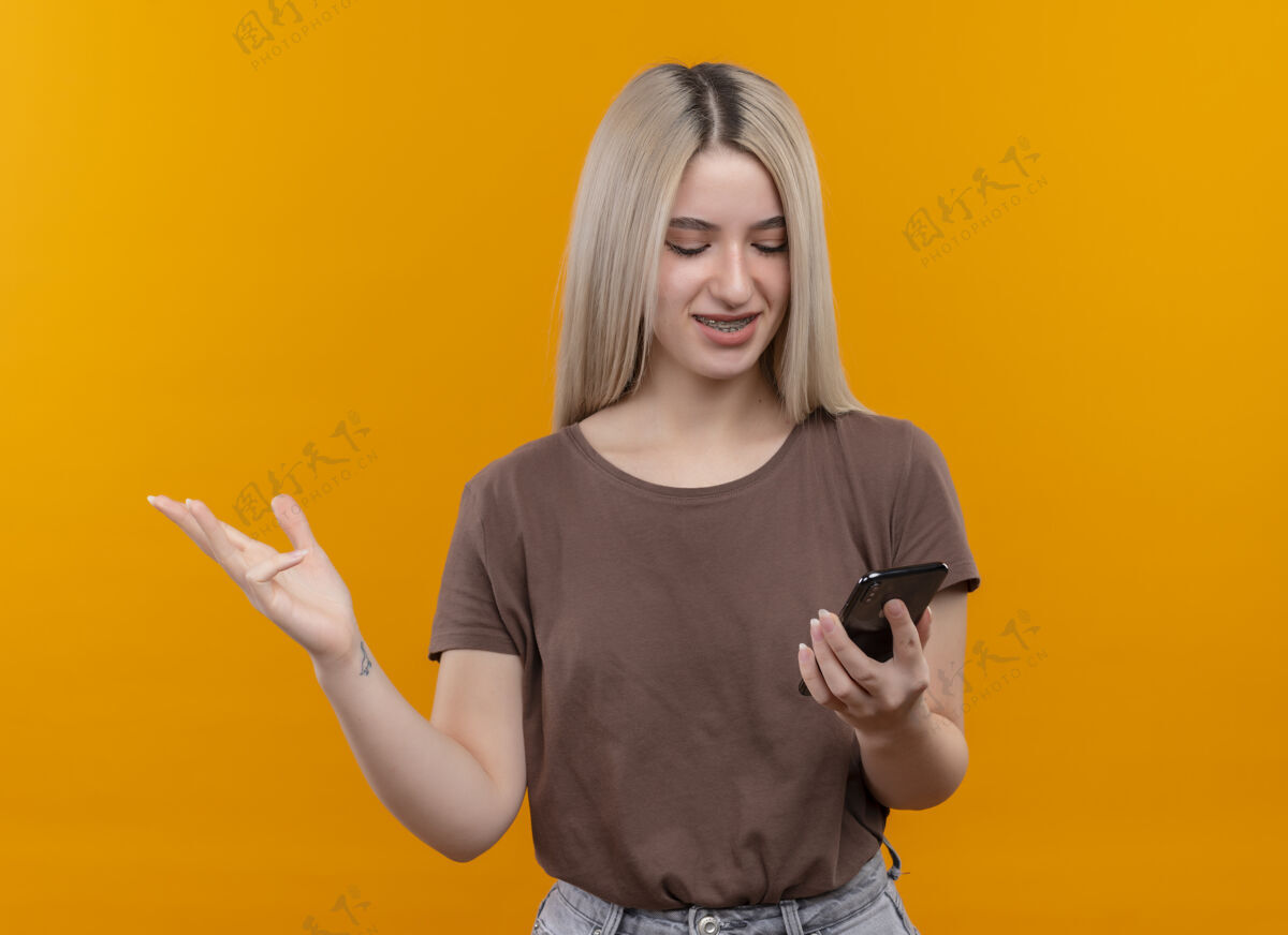 空戴着牙套的金发女孩微笑着拿着手机看着它 在孤立的橙色空间里展示着空手和复印空间金发牙齿女孩