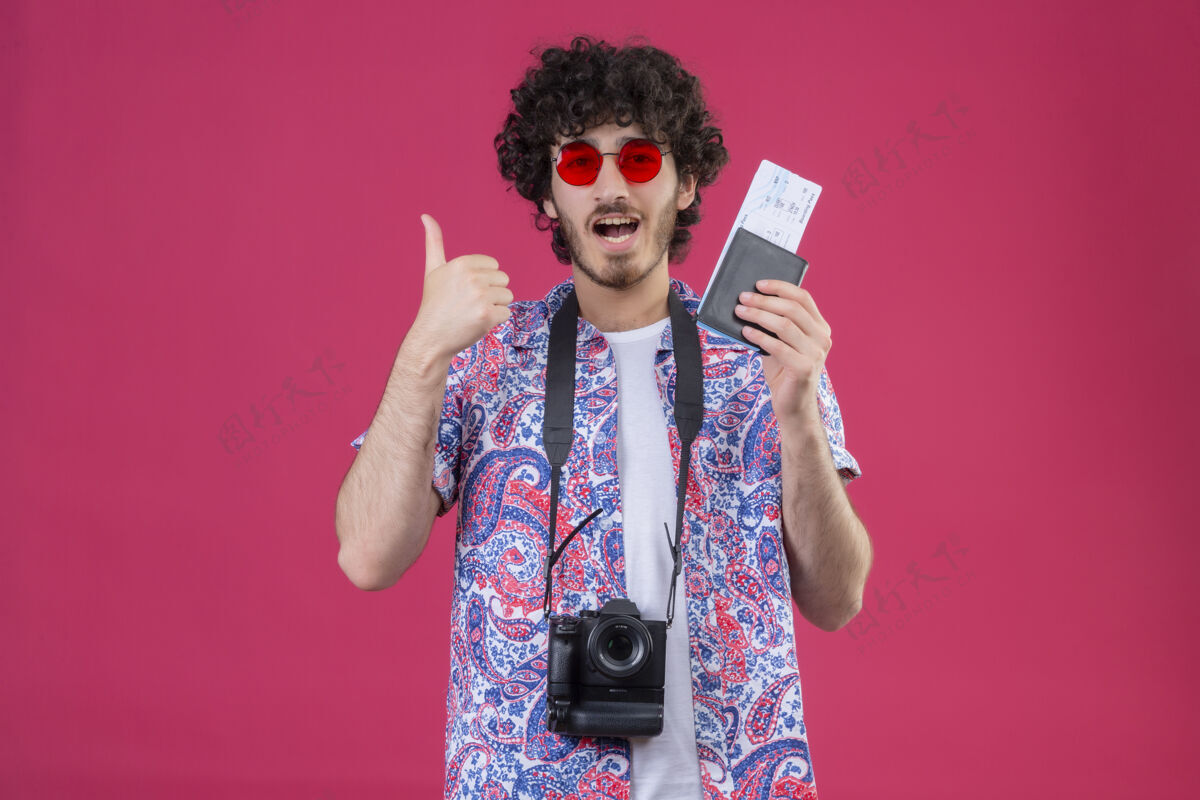 持有快乐的年轻帅气的卷发旅行者戴着太阳镜拿着钱包和机票 脖子上挂着相机 在有复印空间的粉色空间里竖起大拇指欢乐向上穿