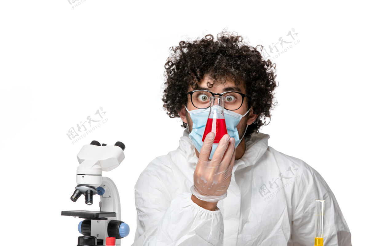 白色正面图：男医生穿着防护服 戴着口罩 拿着装有红色溶液的烧瓶 背景是白色的大流行性冠状病毒病毒视图男医生