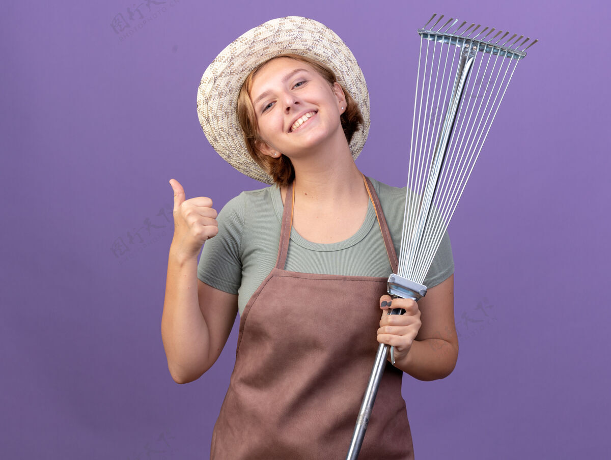 年轻微笑着的年轻斯拉夫女园丁戴着园艺帽竖起大拇指 拿着紫色的叶子耙斯拉夫花园拇指