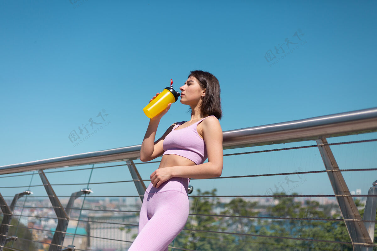 健康年轻女子穿着合身的运动服 在炎热阳光明媚的早晨 在桥上喝着一瓶水 运动后口渴又累蛋白质水锻炼