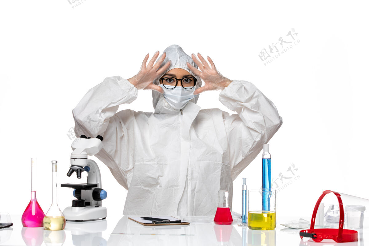 人正面图：穿着特殊防护服的女化学家坐在白色办公桌上 拿着解决方案化学冠状病毒大流行化学桌子医生
