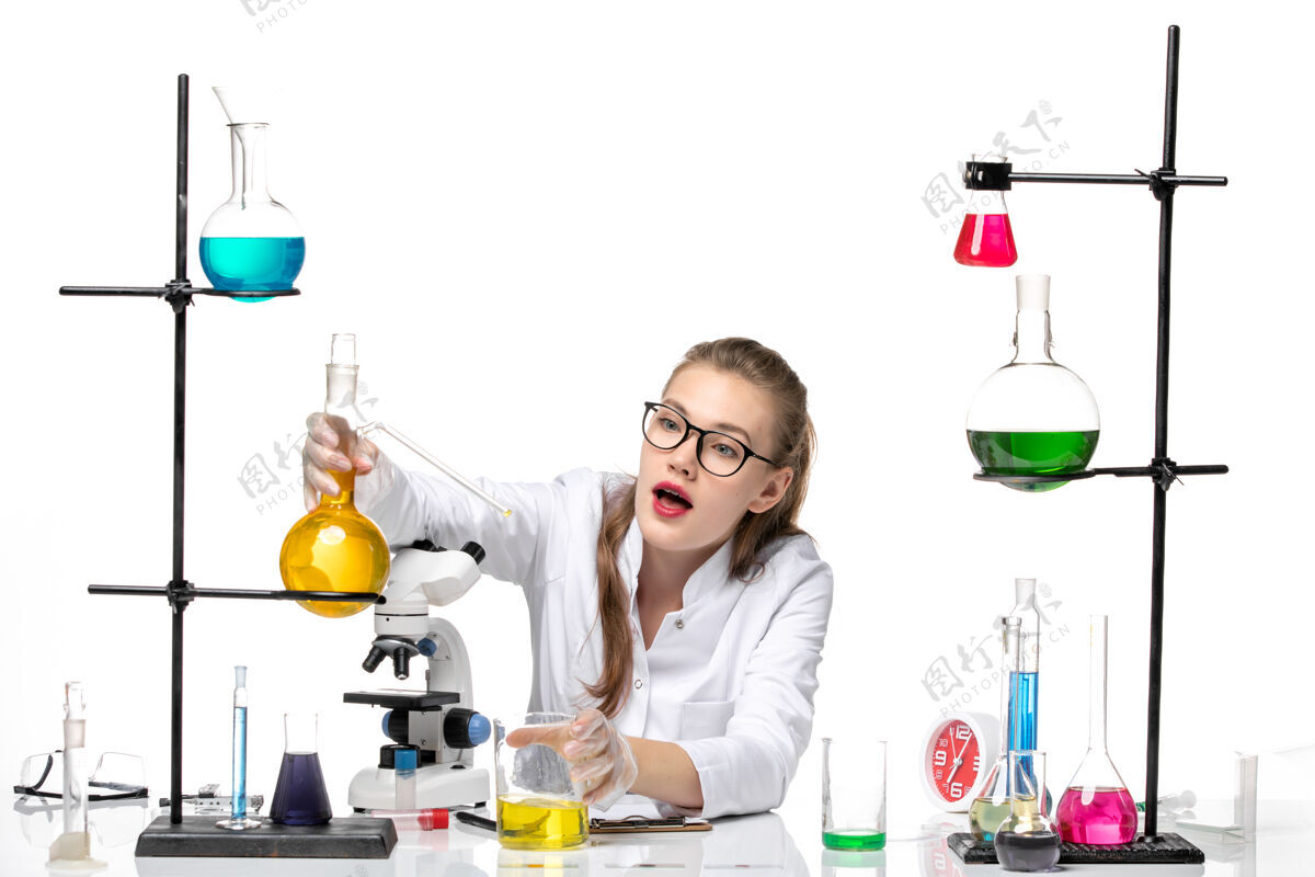 浅白色前视图：身着医疗服的女化学家坐在桌子前 用浅白背景上的溶液化学大流行性冠状病毒医学套装科学