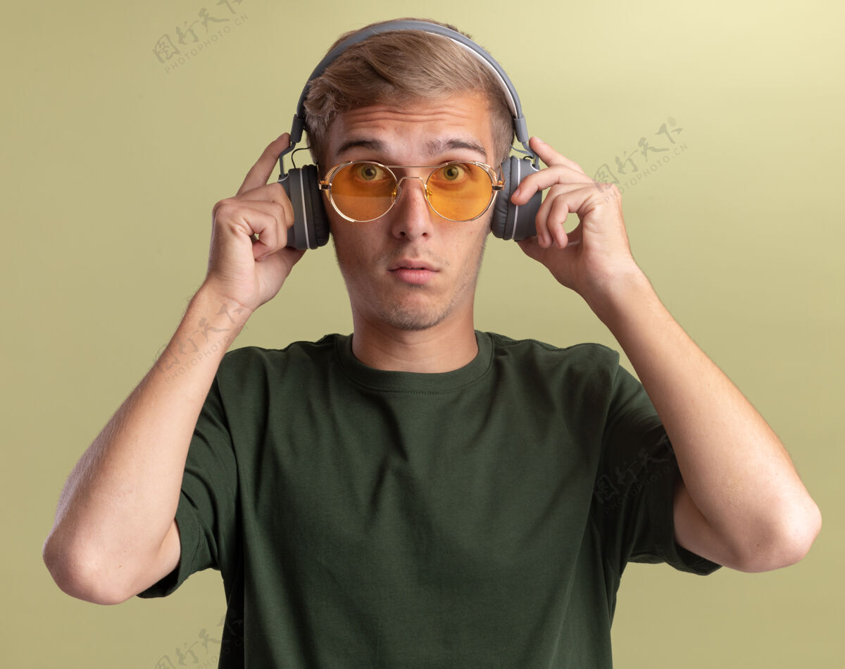 表情惊讶地看着前面的年轻帅哥穿着绿色衬衫 戴着眼镜和耳机隔离在橄榄绿的墙上年轻市民男人