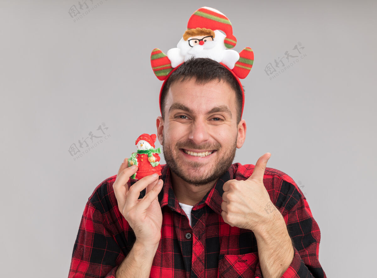 圣诞老人快乐的白人年轻人戴着圣诞老人的头带 手里拿着雪人的圣诞饰品 看着相机 在白色背景上孤立地竖起大拇指显示人圣诞