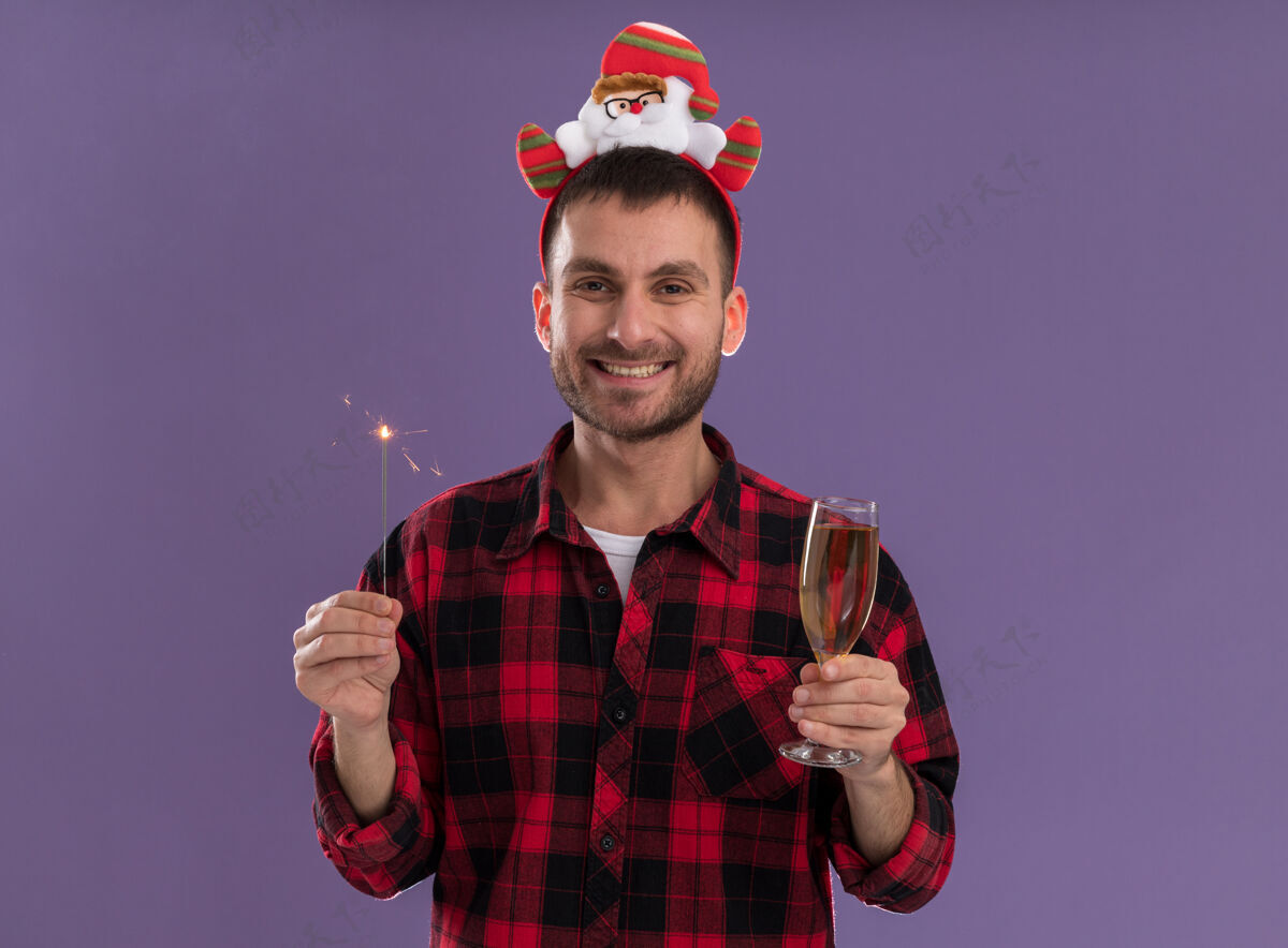 圣诞老人快乐的白人年轻人戴着圣诞老人的头带 手里拿着节日的火花和一杯香槟 看着孤立在紫色背景上的相机快乐头带火花