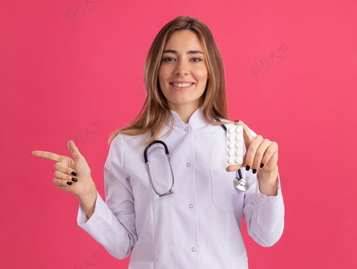 衣服面带微笑的年轻女医生穿着医用长袍 手持听诊器 指着隔离在粉红色墙上的药片长袍听诊器公民