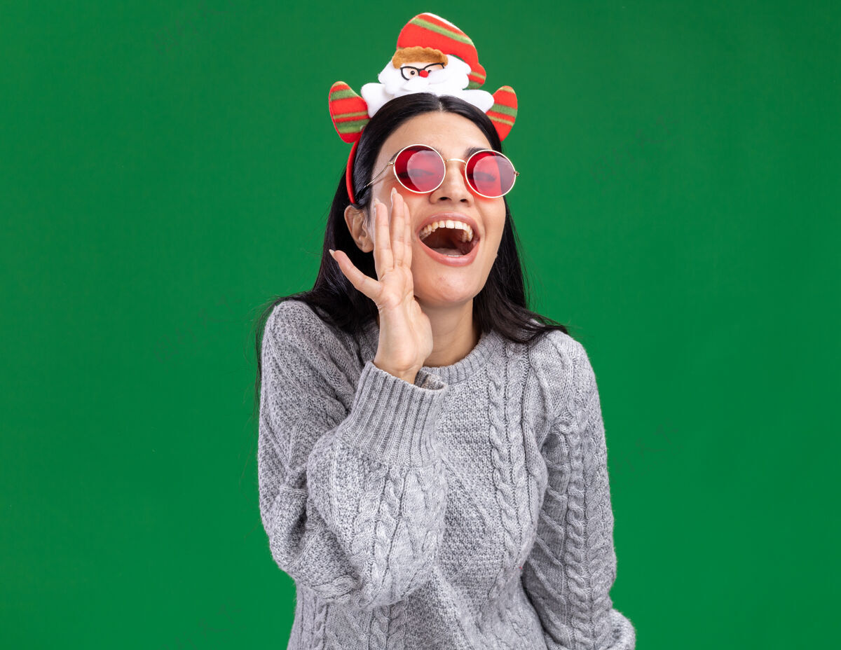 耳语快乐的年轻白人女孩戴着圣诞老人的头带 戴着眼镜 看着相机 在绿色的背景上低语圣诞老人圣诞老人圣诞快乐