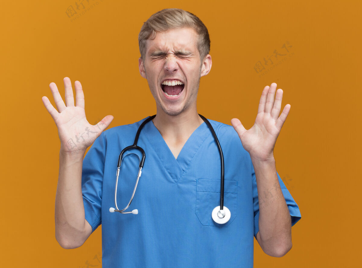 穿着兴奋的闭着眼睛年轻的男医生穿着医生制服听诊器摊开双手隔离在橙色的墙上眼睛衣服人