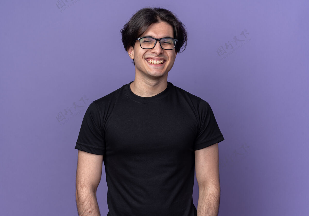 姿势笑容可掬的年轻帅哥 穿着黑色t恤 戴着眼镜孤立在紫色的墙上脸衣服市民