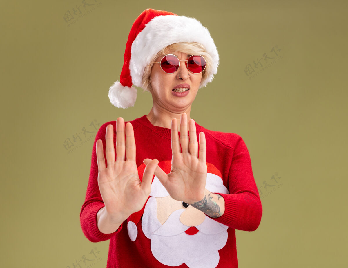 圣诞快乐恼怒的金发女郎戴着圣诞帽 穿着圣诞老人的圣诞毛衣 戴着眼镜 在橄榄绿的背景下看着摄像机做着拒绝的手势圣诞节圣诞老人复制