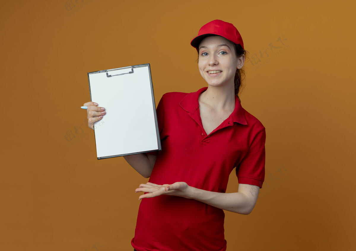 帽子微笑着的年轻漂亮的送货女孩 穿着红色制服 戴着帽子 手里拿着笔 拿着剪贴板 用手指着孤立在橙色背景上的剪贴板 还有复印空间手漂亮女孩