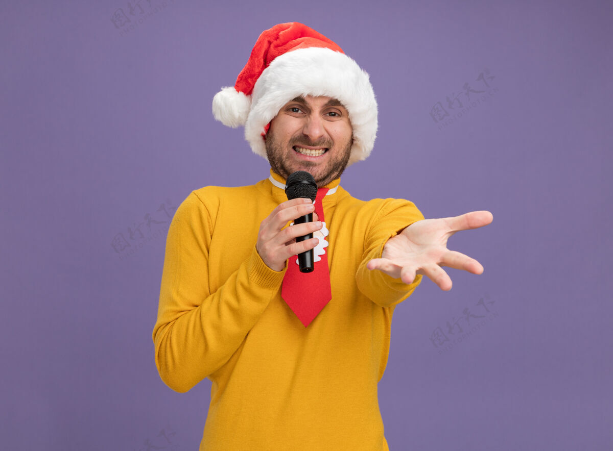 帽子恼怒的年轻白人男子戴着圣诞帽打着领带拿着麦克风看着相机伸出手对着隔离在紫色背景上的相机空间男人走向
