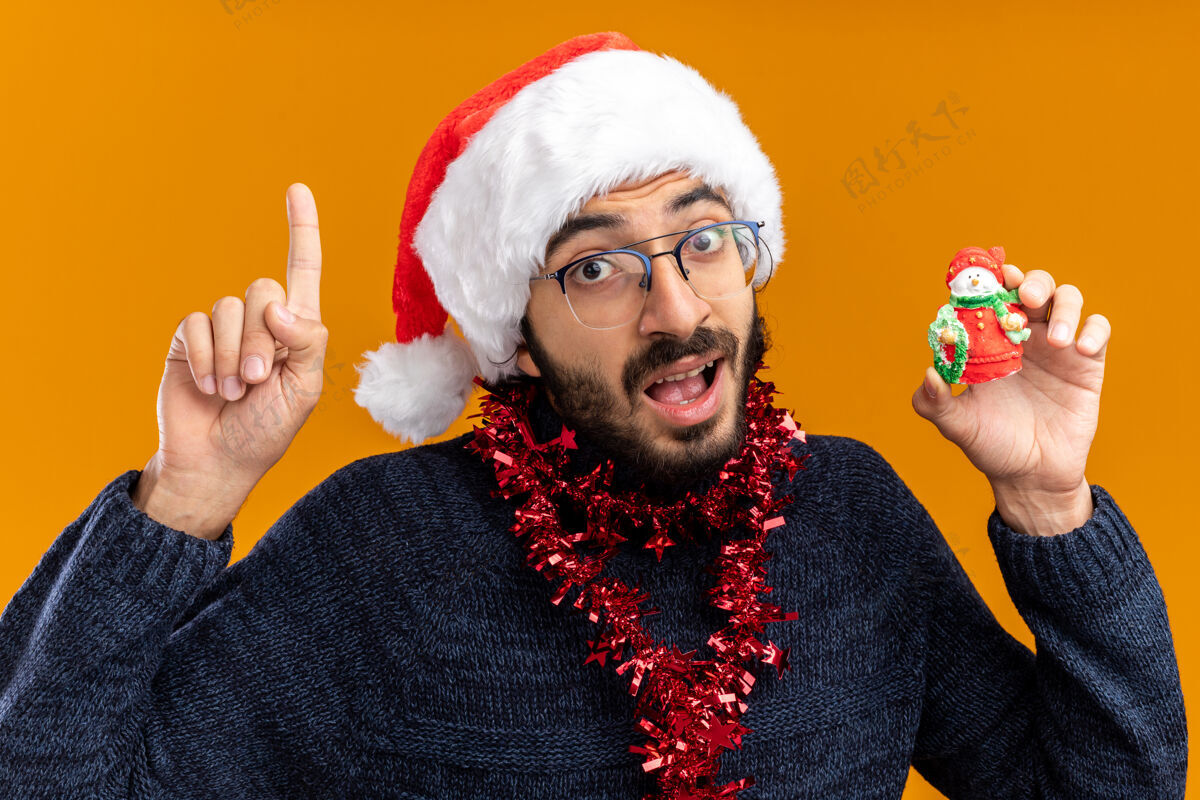 家伙给人印象深刻的年轻帅哥戴着圣诞帽 脖子上戴着花环 拿着玩具点在橙色背景上孤立起来印象年轻圣诞快乐