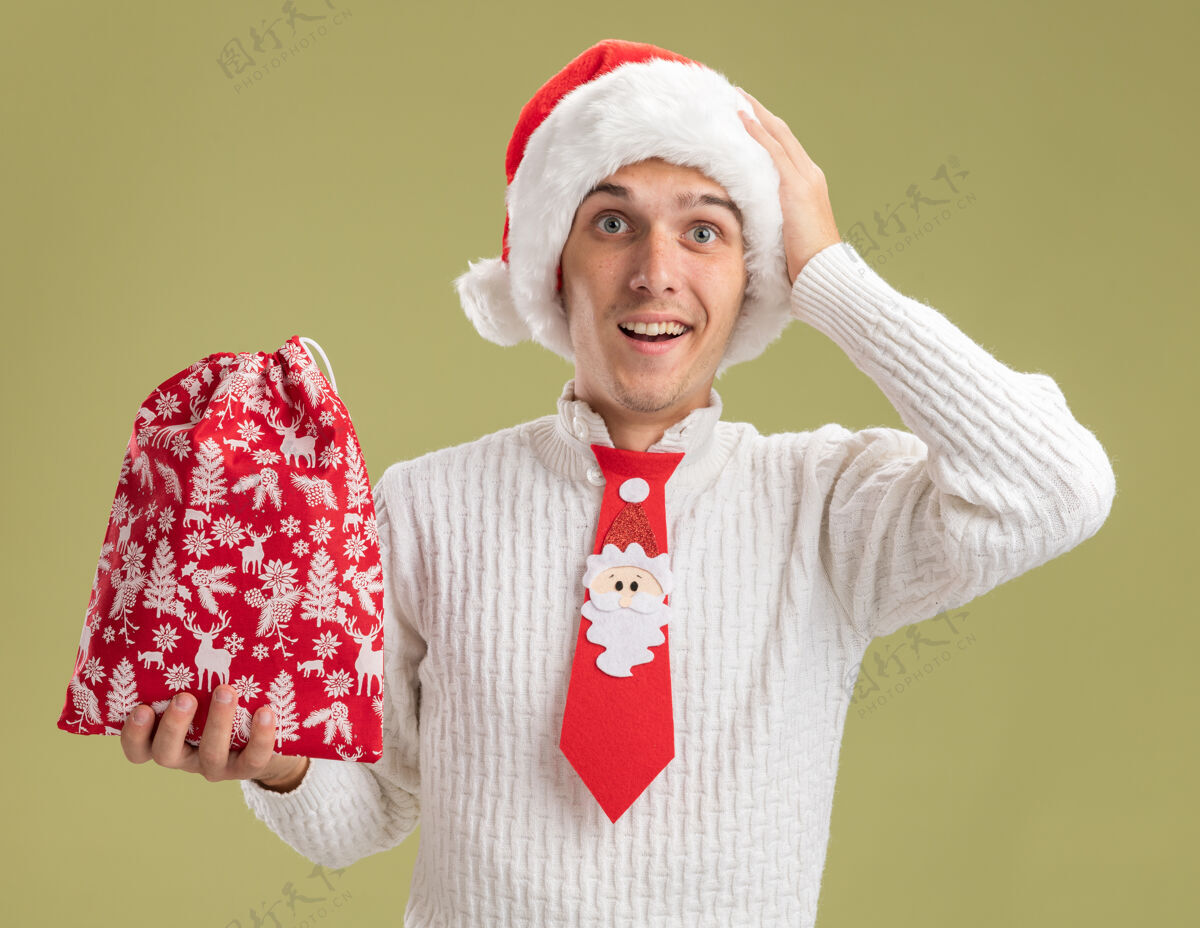 抱着印象深刻的年轻帅哥戴着圣诞帽 打着圣诞老人的领带 手里拿着圣诞袋 手放在头上 看着橄榄绿背景上的相机圣诞手袋子