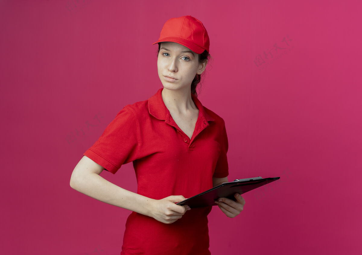 女孩自信的年轻漂亮的送货女孩 穿着红色制服 戴着帽子 拿着剪贴板 看着摄像机 被隔离在深红色的背景上 还有复印空间自信漂亮剪贴板