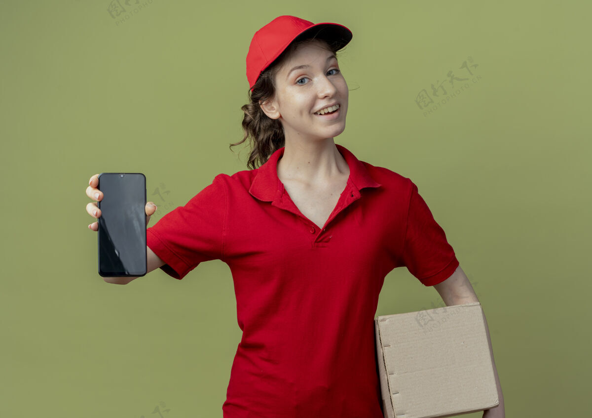女孩快乐的年轻漂亮的送货女孩 穿着红色制服 戴着帽子 拿着纸箱 在橄榄绿的背景下 把手机伸向镜头伸展快乐帽子
