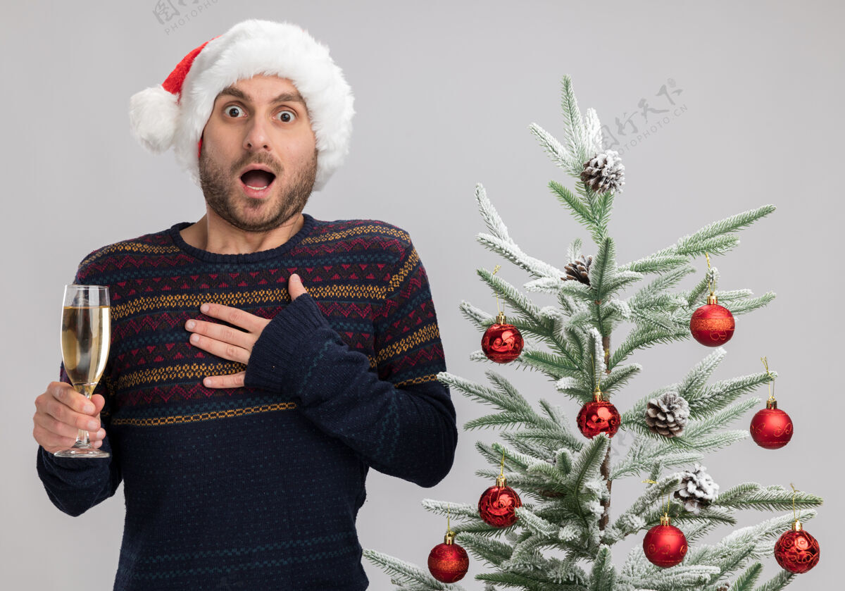 香槟令人印象深刻的年轻白种人戴着圣诞帽站在圣诞树旁拿着一杯香槟看着相机手放在胸前隔离在白色背景上印象圣诞圣诞快乐
