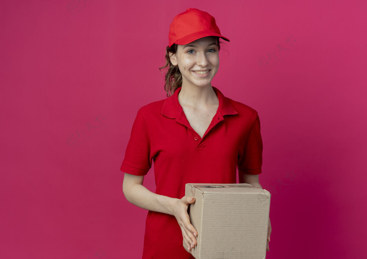 持有微笑着的年轻漂亮的送货女孩 穿着红色制服 戴着帽子 拿着纸箱 在深红色的背景上与复印空间隔离开来红色女孩帽子