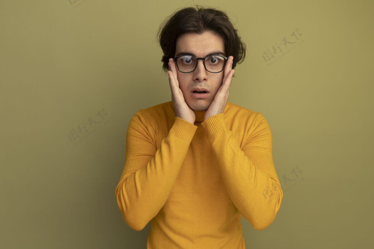 姿势惊讶的年轻帅哥穿着黄色高领毛衣 戴着眼镜 双手放在橄榄绿的墙上孤立的脸颊上帅哥人穿着