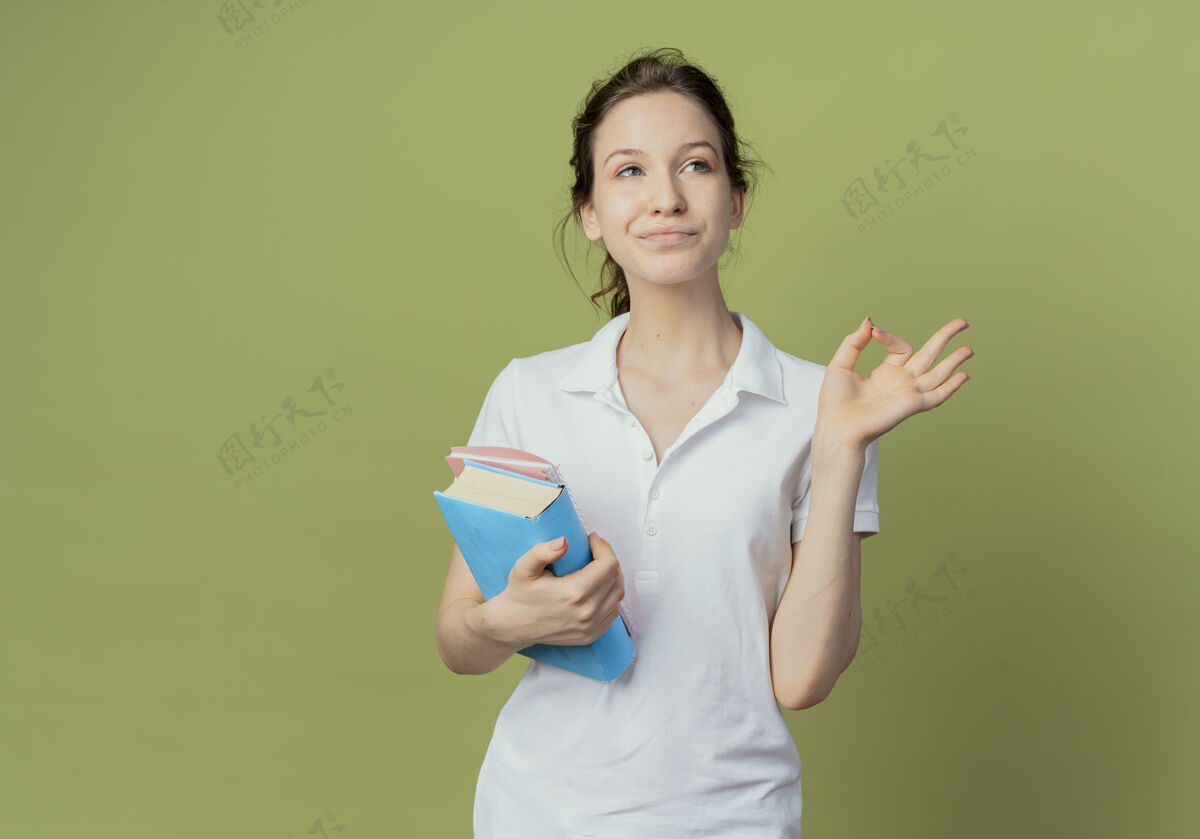 橄榄色高兴的年轻漂亮的女学生拿着书和便笺簿看在一边 做着ok的标志在橄榄绿的背景上与复印空间隔离垫壁板注意