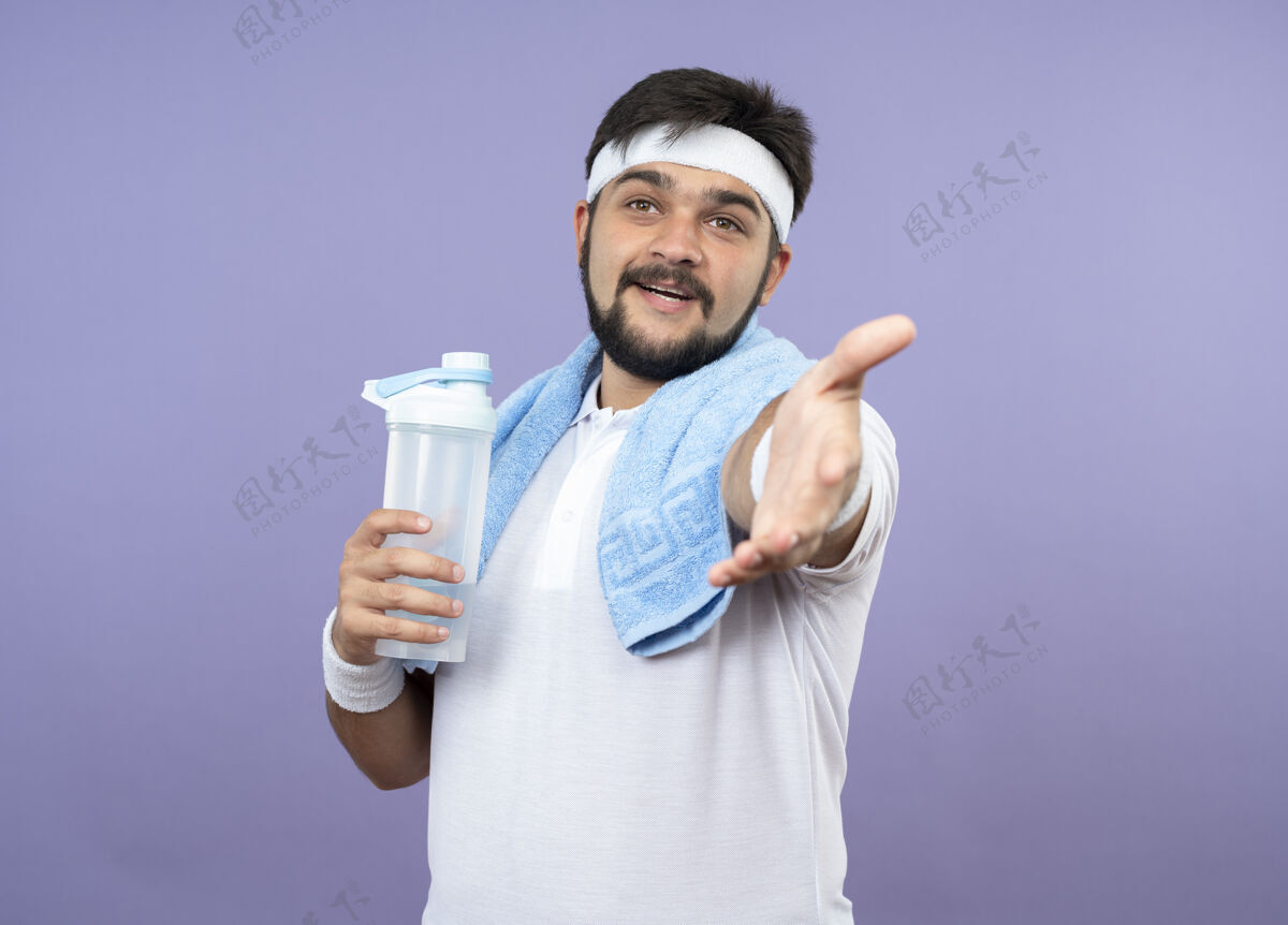运动型高兴的年轻人戴着头带和腕带拿着水瓶与毛巾在肩上伸出手毛巾瓶子人