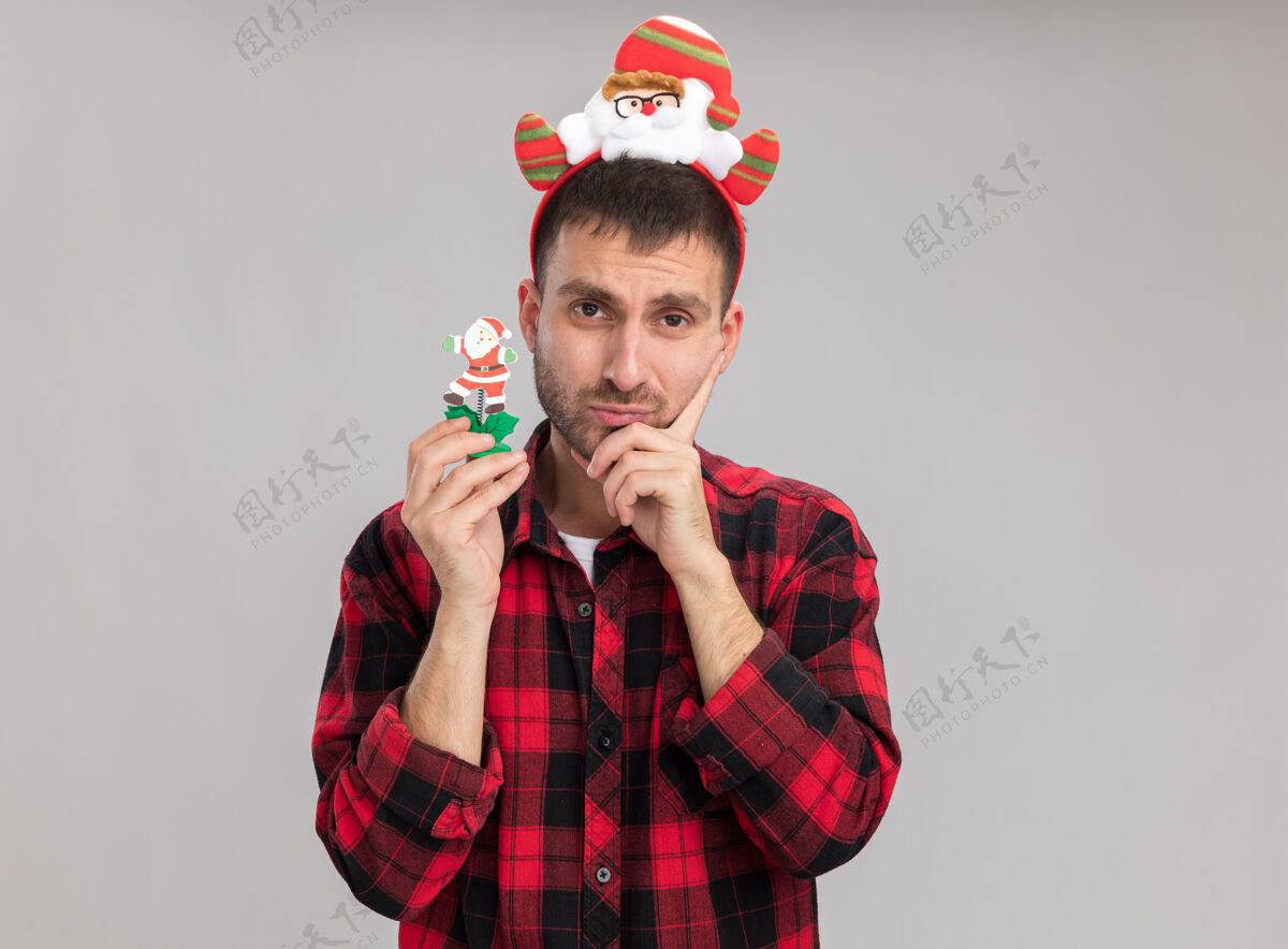 头带困惑的白人年轻人戴着圣诞老人的头带拿着雪人的圣诞玩具看着相机手放在下巴上隔离在白色背景上复制玩具困惑