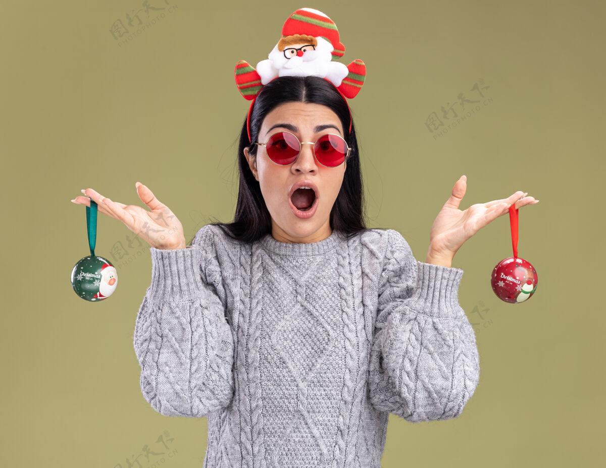 抱着令人印象深刻的年轻白人女孩戴着圣诞老人的头带 戴着眼镜 拿着圣诞饰品 看着橄榄绿背景上的相机圣诞饰品圣诞老人年轻