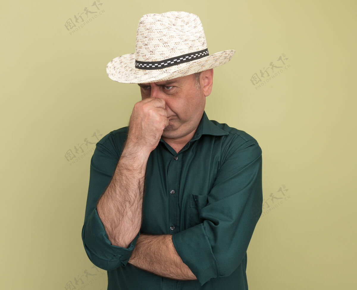 表情不高兴地看着身边穿着绿色t恤和帽子的中年男子抓住了孤立在橄榄绿墙上的鼻子感觉站脸