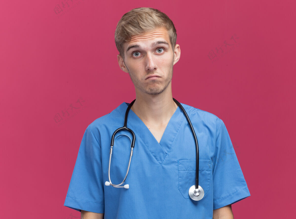 男性年轻的男医生穿着带听诊器的医生制服-隔离在粉红色的墙上 留有复印空间相机表情脸