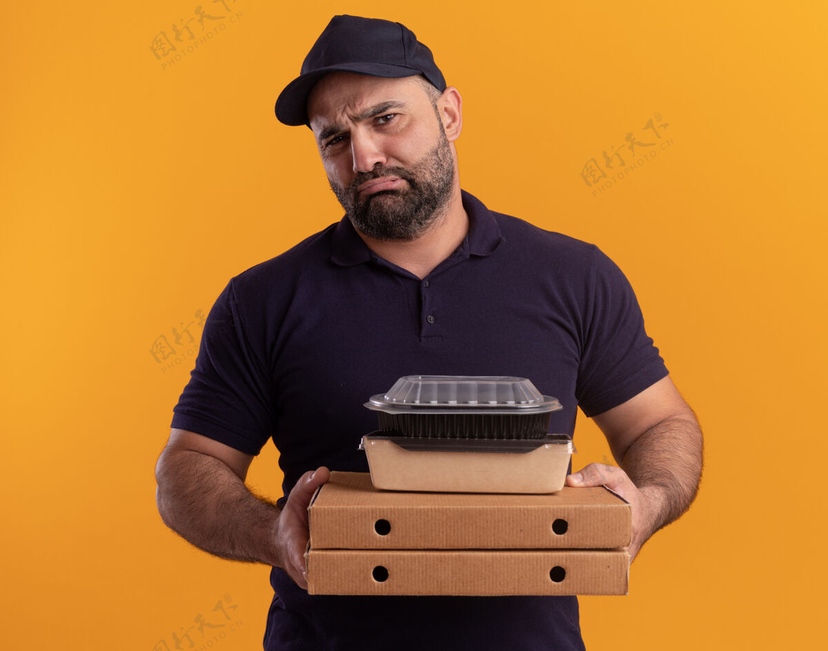 容器悲伤的中年送货员穿着制服 戴着帽子 拿着食物容器放在隔离在黄色墙上的比萨饼盒上制服衣服披萨