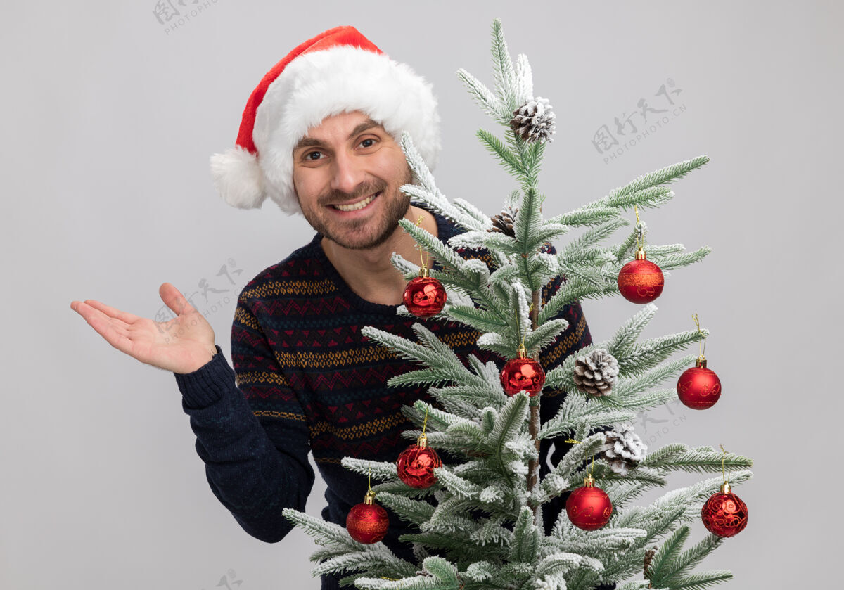 白快乐的年轻白人男子戴着圣诞帽站在圣诞树后 看着相机显示空手孤立在白色背景圣诞表演圣诞快乐
