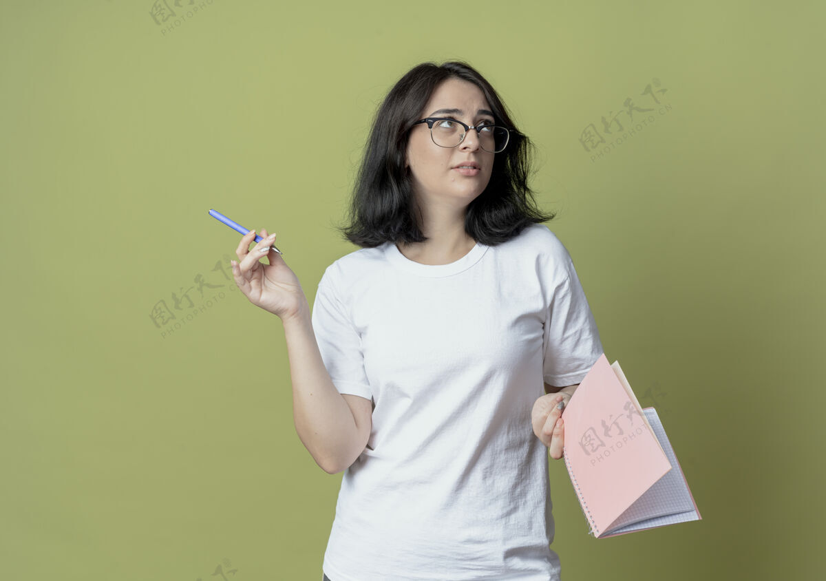 垫体贴的年轻漂亮的高加索女孩戴着眼镜拿着笔和笔记本看着一边孤立的橄榄绿背景和复印空间笔记拿着壁板