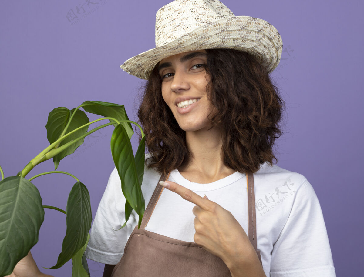 园艺年轻的女园丁面带微笑 穿着制服 戴着园艺帽 指着植物生活方式快乐点